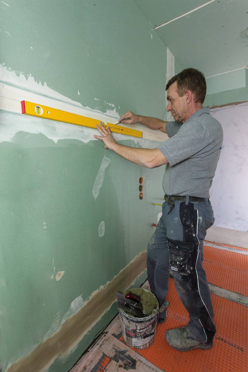 Ein Handwerker im grauen T-Shirt richtet eine Wasserwaage an einer grünen Wand aus, während er an einem Innenausbauprojekt arbeitet.