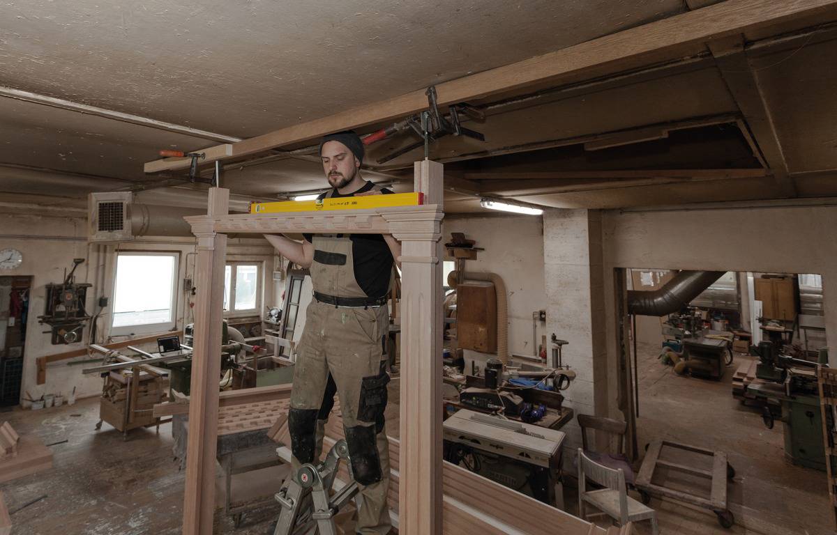 Ein Handwerker in einer Werkstatt steht auf einer Leiter und verwendet eine Wasserwaage, um einen Holzbalken zu messen. Im Hintergrund sind Maschinen und Werkzeuge sichtbar.