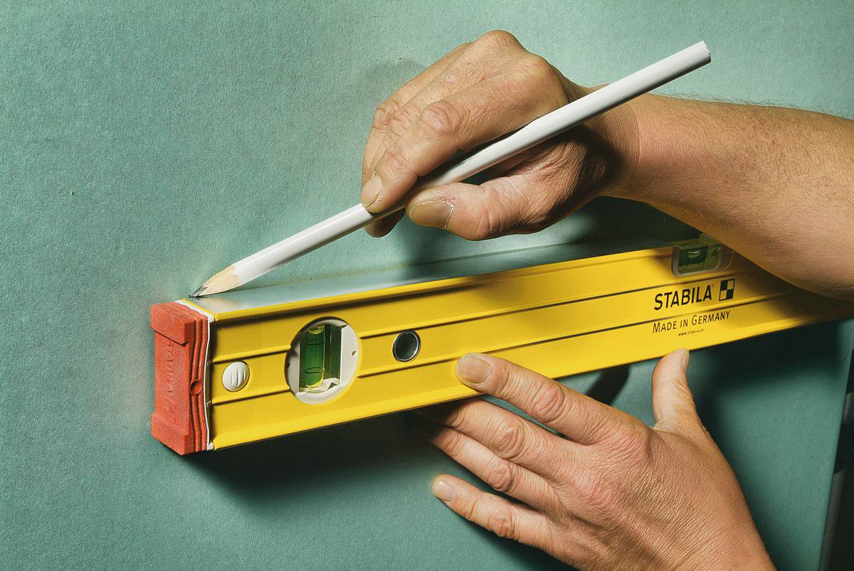 Eine Hand hält eine Wasserwaage an einer Wand, während die andere Hand mit einem Bleistift Markierungen macht.