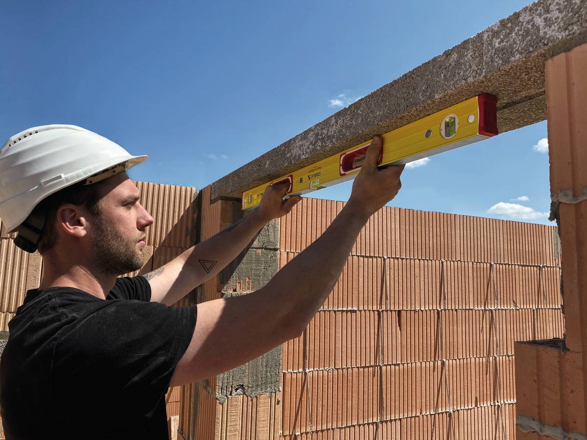 Ein Bauarbeiter mit Schutzhelm nutzt eine Wasserwaage, um eine Mauer aus roten Ziegeln zu prüfen. Der Himmel ist blau und wolkenlos.