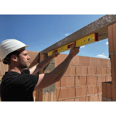 Ein Bauarbeiter mit Schutzhelm nutzt eine Wasserwaage, um eine Mauer aus roten Ziegeln zu prüfen. Der Himmel ist blau und wolkenlos.