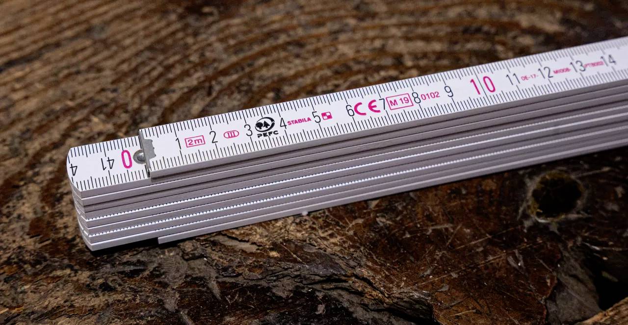 Ein weißer Zollstock mit roten Markierungen liegt auf einem Holzstamm. Die Maßskala zeigt bis 10 Zentimeter.