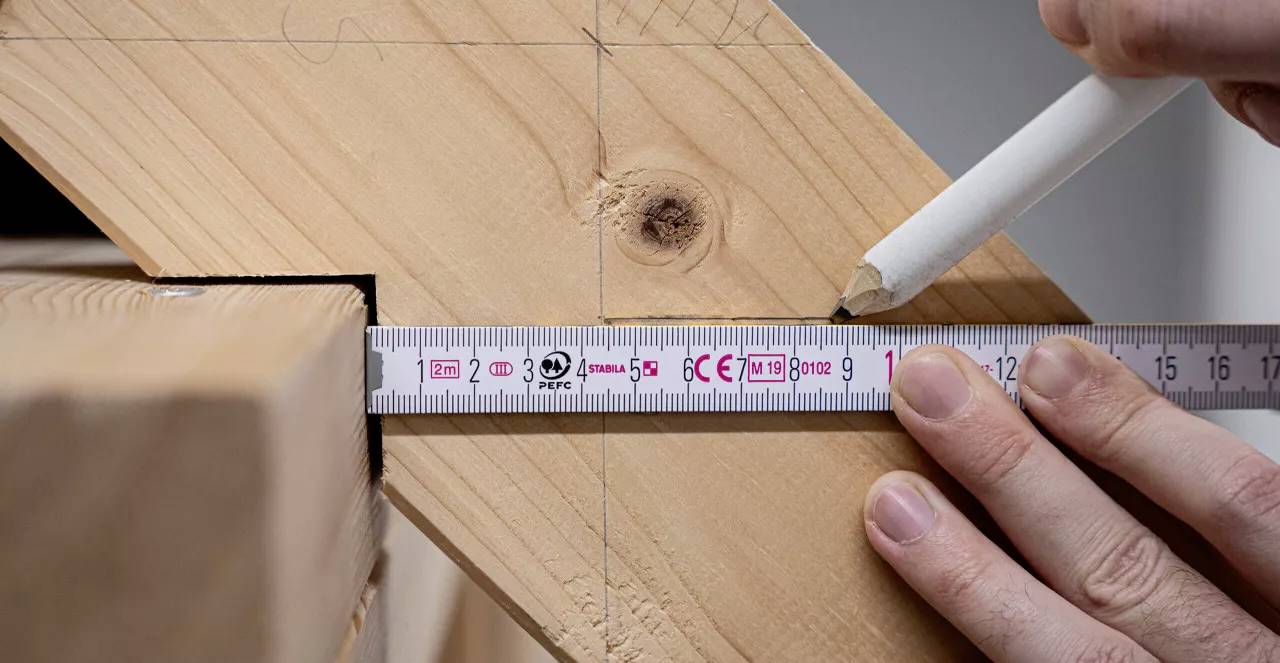 Ein Lineal liegt auf einem Holzbalken, während eine Hand mit einem weißen Bleistift eine Markierung an einem gemessenen Punkt setzt.