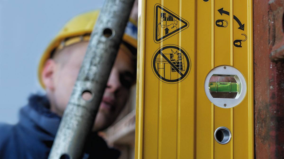 Eine Person mit Helm hält eine Leiter, daneben eine Wasserwaage mit Symbolen zur sicheren Nutzung. Fokus auf Sicherheit am Arbeitsplatz.