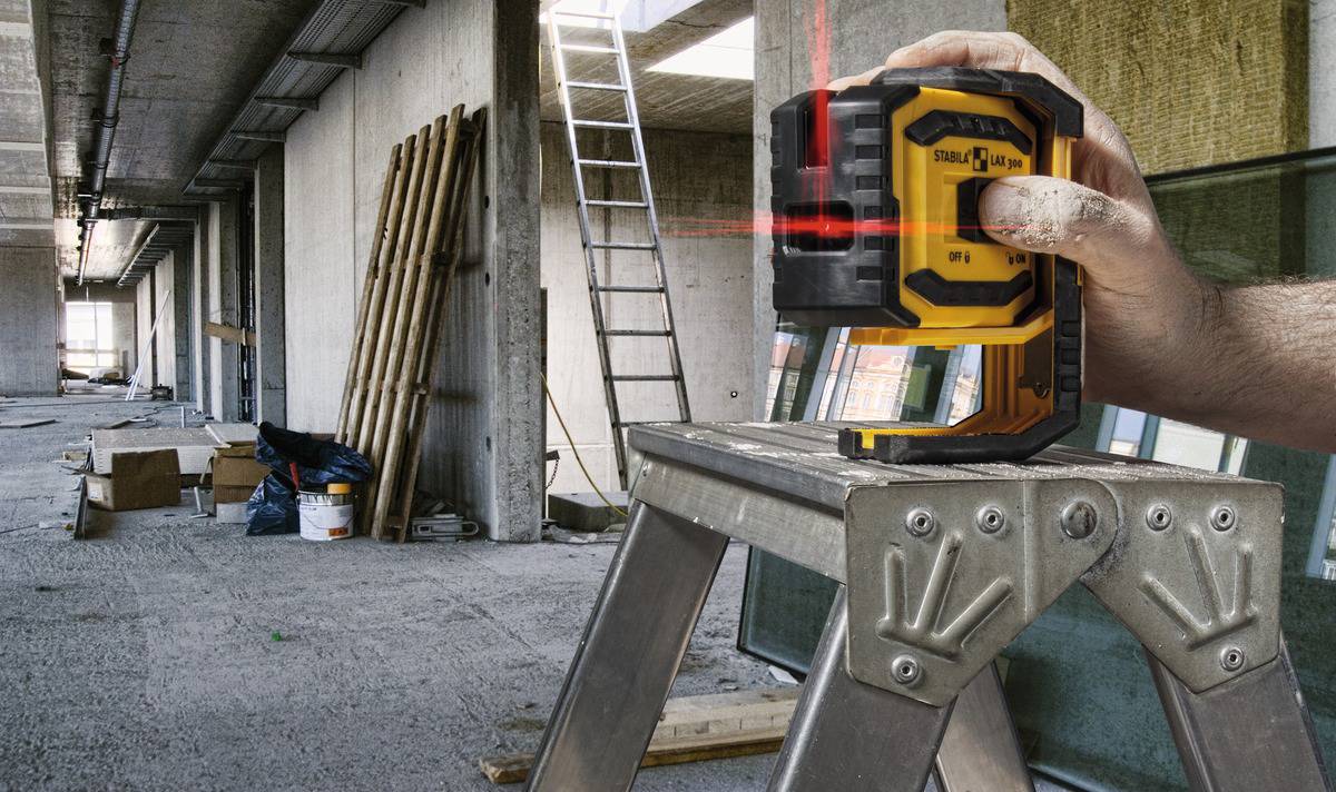 Eine Hand hält ein Lasernivelliergerät auf einer Leiter in einem unfertigen Gebäude. Baustelle mit Bauzubehör im Hintergrund.