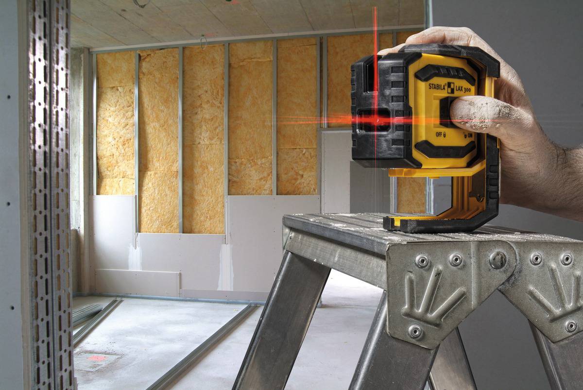 Ein Handwerker benutzt einen gelben Laser-Entfernungsmesser auf einer Leiter, um eine Wand in einem im Bau befindlichen Raum auszurichten.