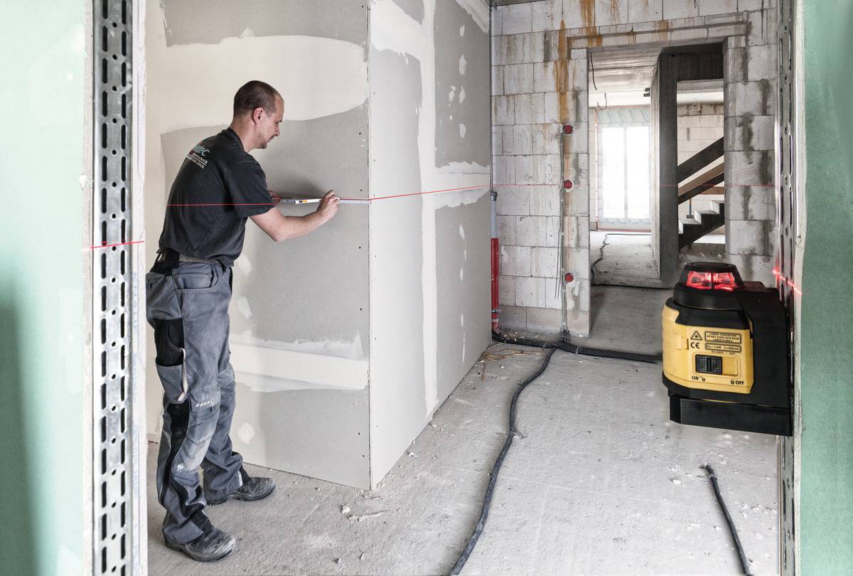 Ein Bauarbeiter misst mit einem roten Laser Entfernung an einer Trockenbauwand in einem unfertigen Raum. Ein gelbes Gerät steht daneben.