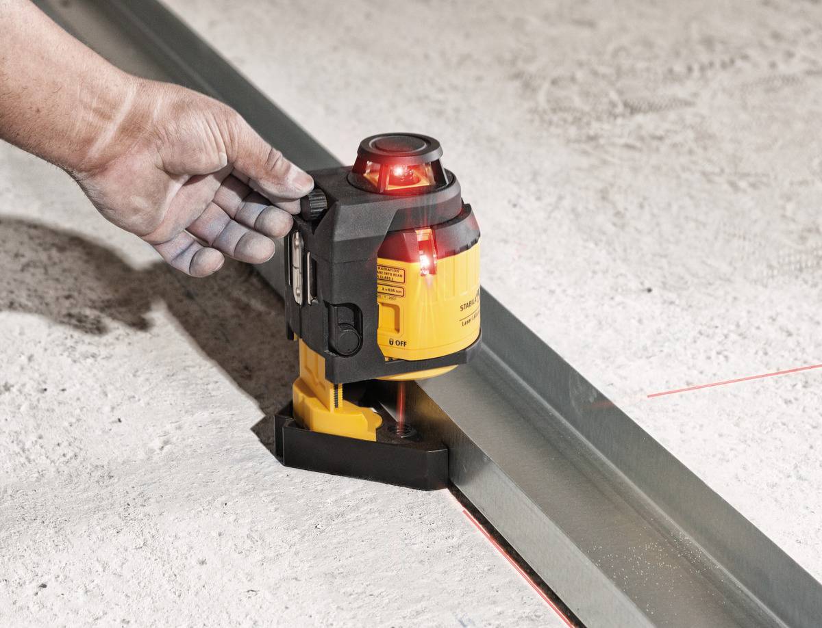 Eine Hand hält ein gelbes Lasergerät, das horizontale rote Laserlinien entlang einer Metallschiene auf einem Betonboden projiziert.
