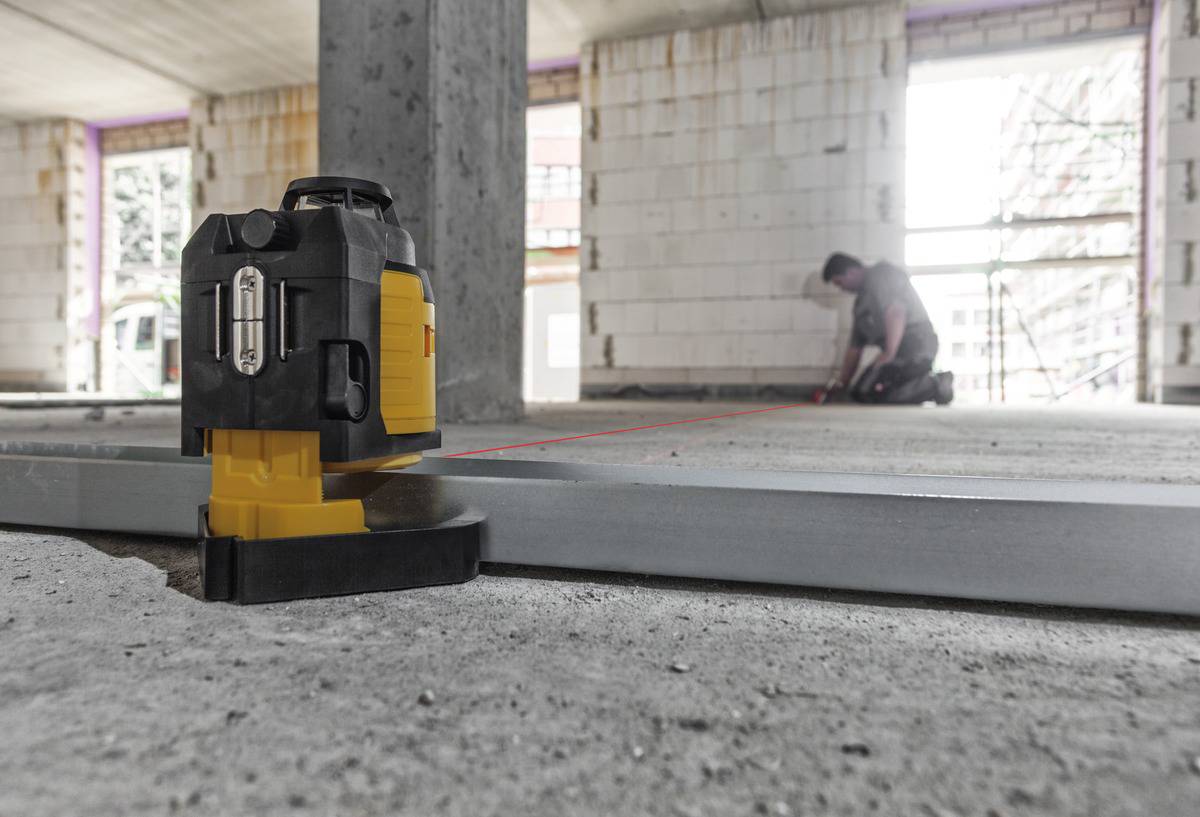 Ein rotierender Laser auf einer Baustelle projiziert eine Linie auf den Boden, während ein Arbeiter im Hintergrund Messungen vornimmt.