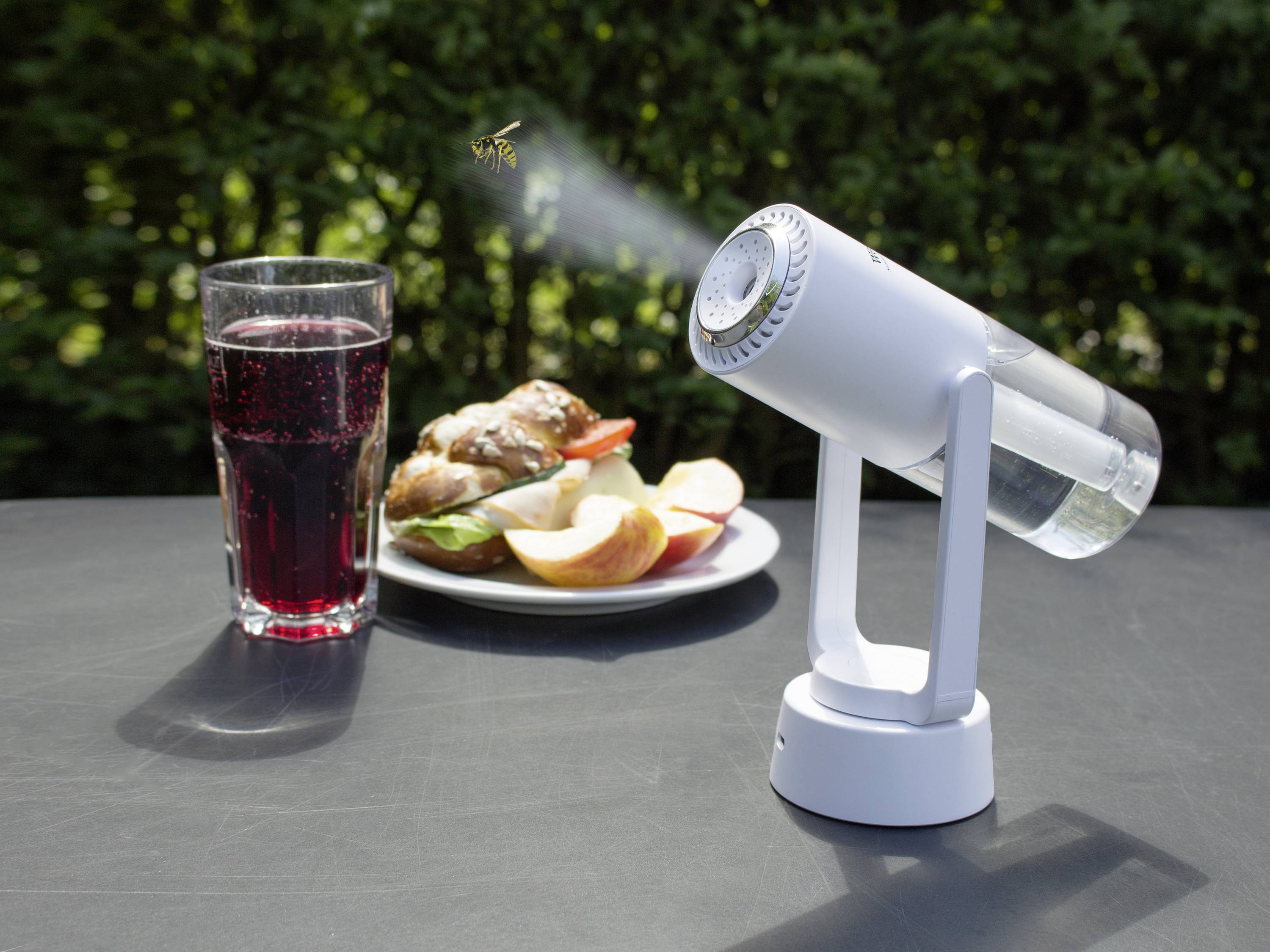 Kleiner Ventilator stößt Dunst aus, während eine Wespe in der Nähe eines Tellers mit Essen und einem Glas roter Flüssigkeit im Freien fliegt.