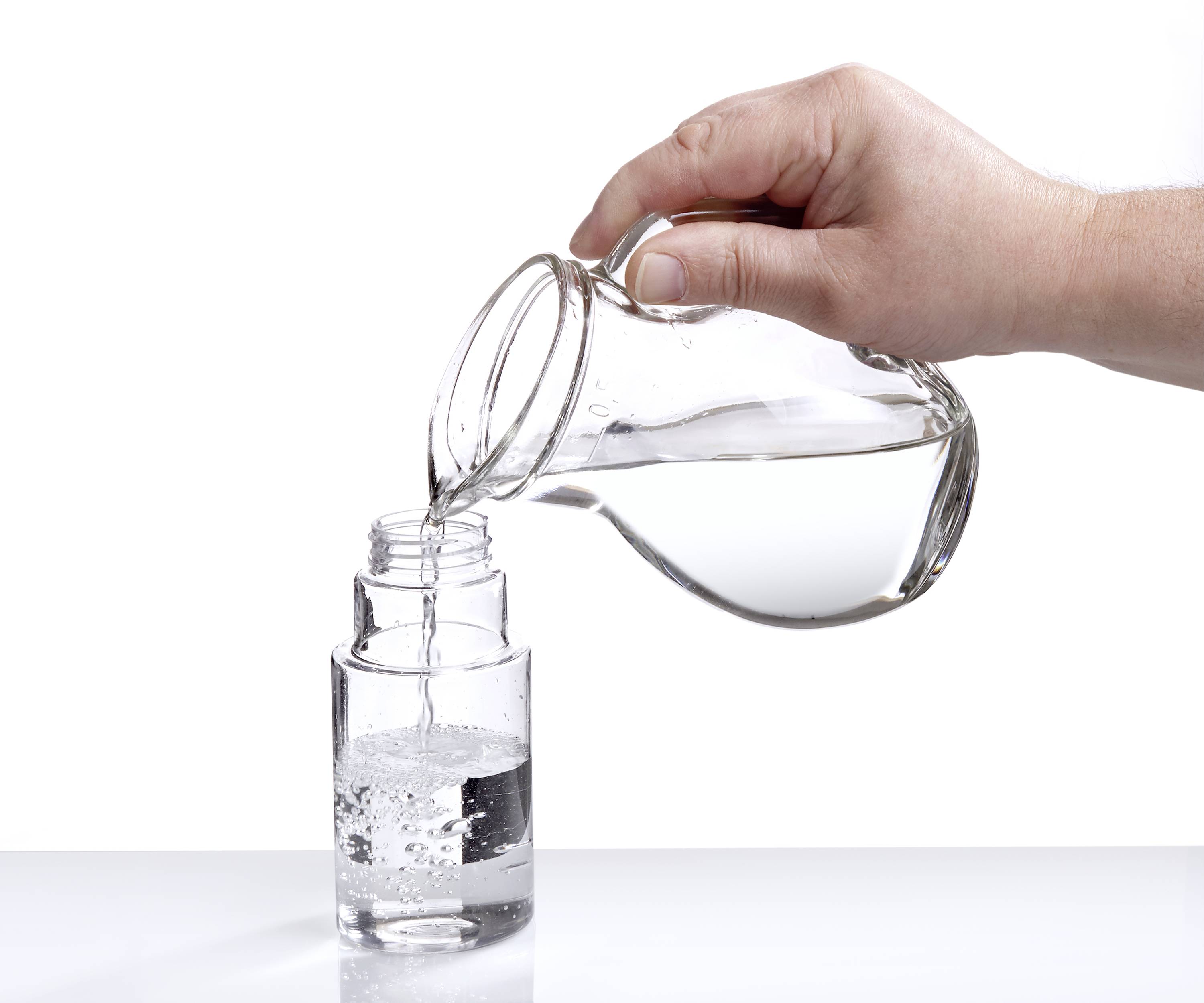 Eine Hand gießt Wasser von einem Glaskrug in ein Glas, das zur Hälfte gefüllt auf einer weißen Oberfläche steht.