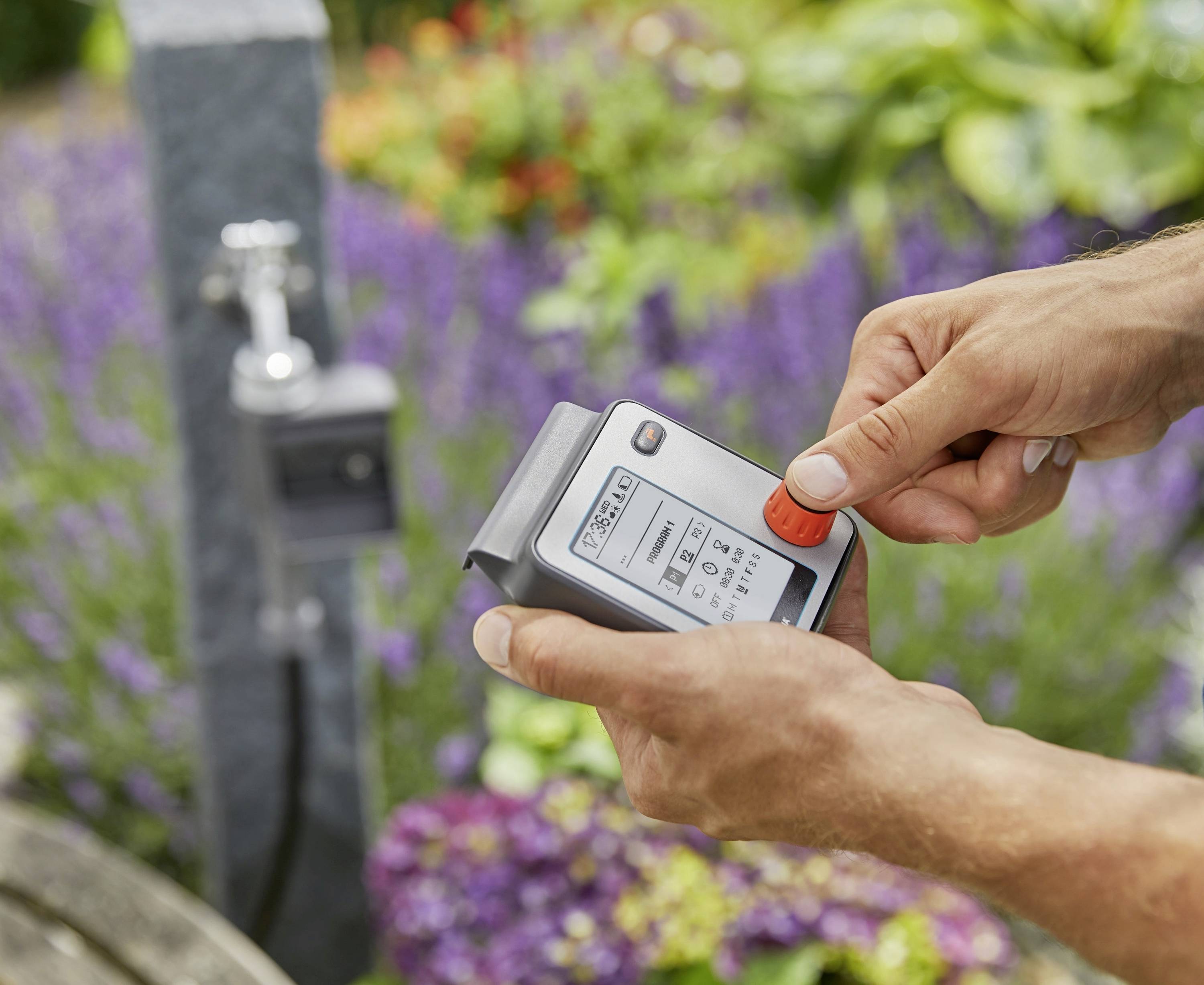 Eine Person hält ein digitales Bewässerungsgerät vor einer Gartenkulisse mit unscharfen Blumen.
