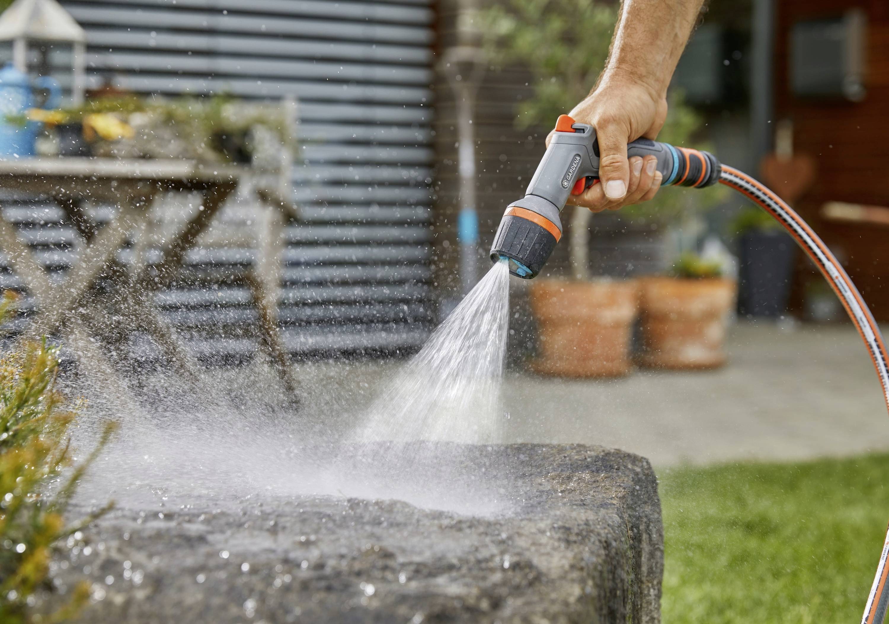 Eine Person gießt mit einer Sprühdüse Wasser auf einen großen flachen Stein im Garten. Im Hintergrund sind Pflanztöpfe zu sehen.