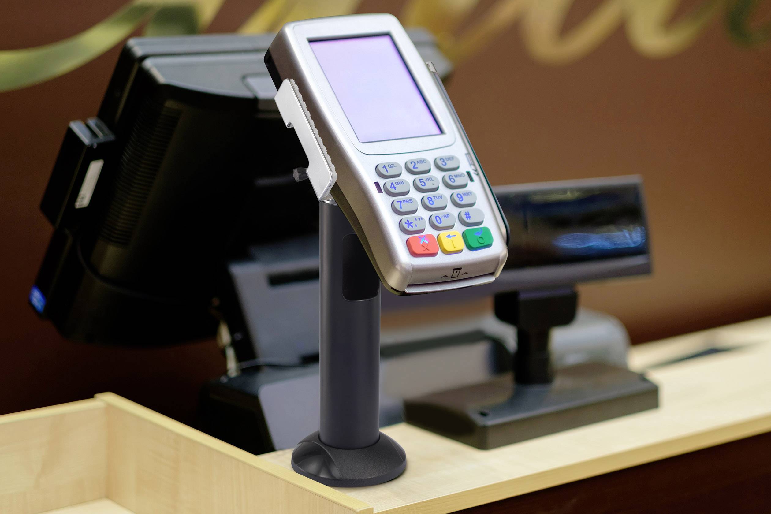 Ein Kartenterminal auf einem Ständer befindet sich auf einer Ladenkasse, bereit für Zahlungen. Im Hintergrund ist eine Registrierkasse sichtbar.