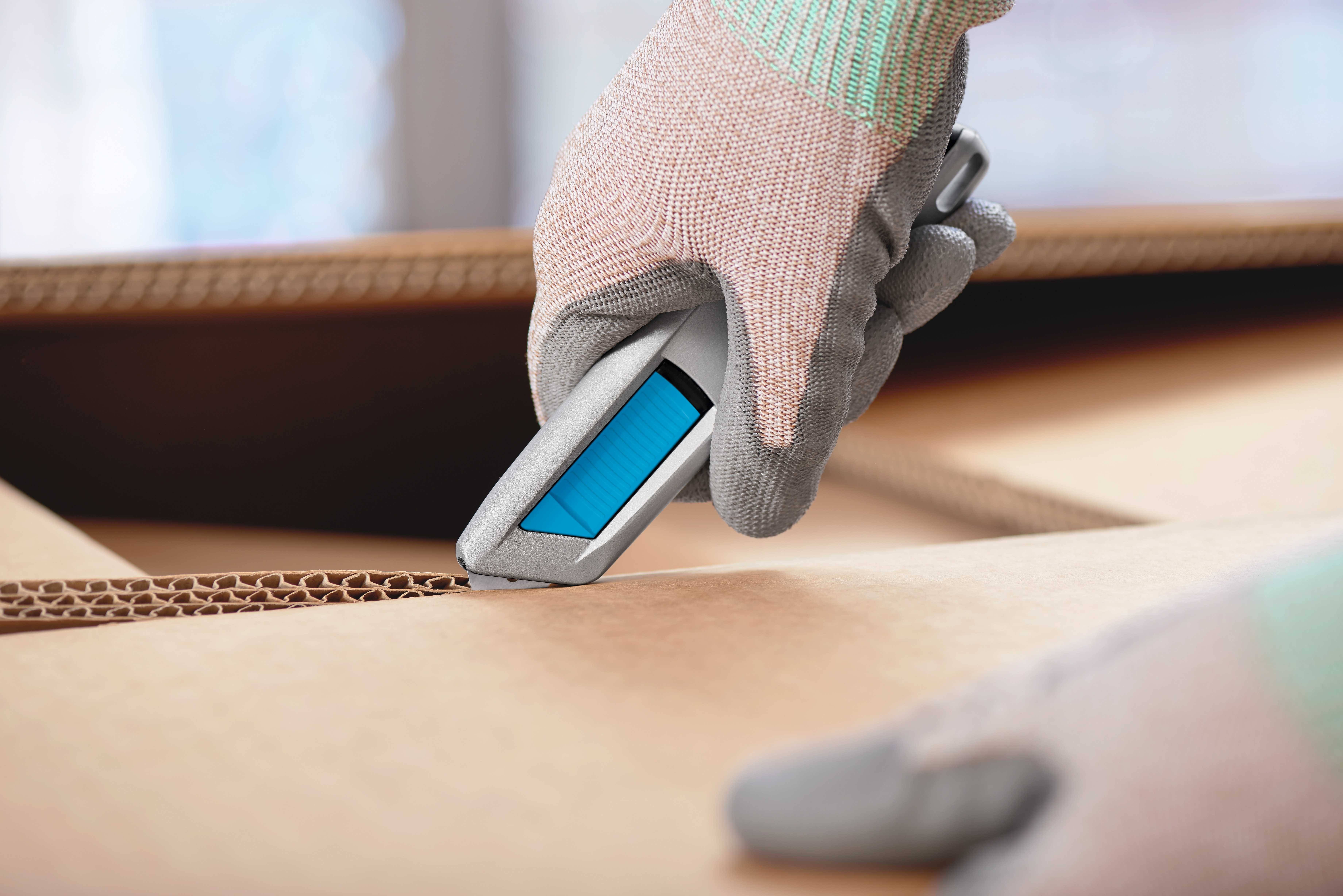 'Nahaufnahme einer Hand mit Handschuh, die einen Karton mit einem blauen Cutter-Messer öffnet. Die Szene zeigt Sorgfalt bei der Arbeit.'