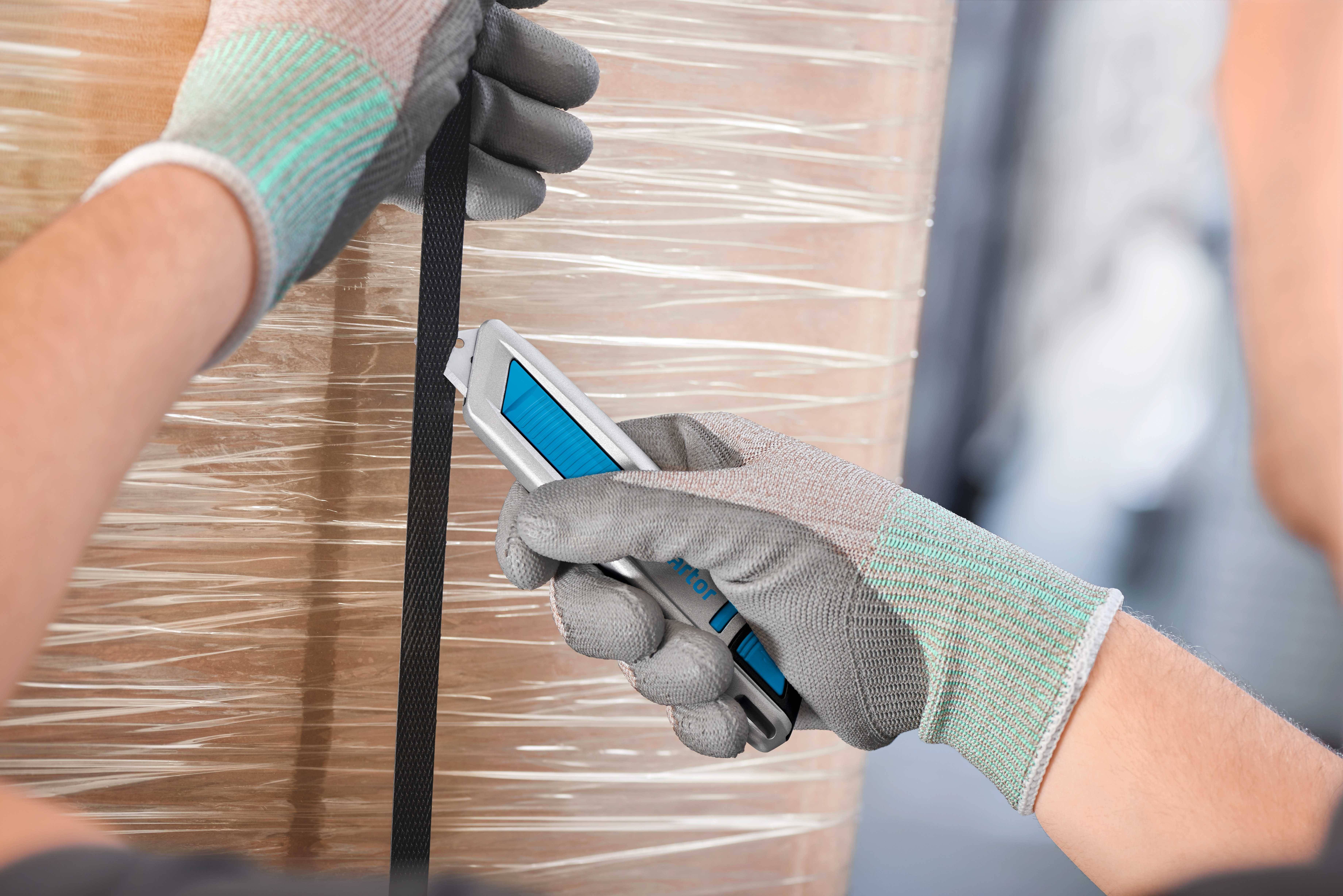 Eine Person schneidet mit einem Cuttermesser ein schwarzes Band, während sie Schutzhandschuhe trägt. Im Hintergrund ist eine verpackte Palette.