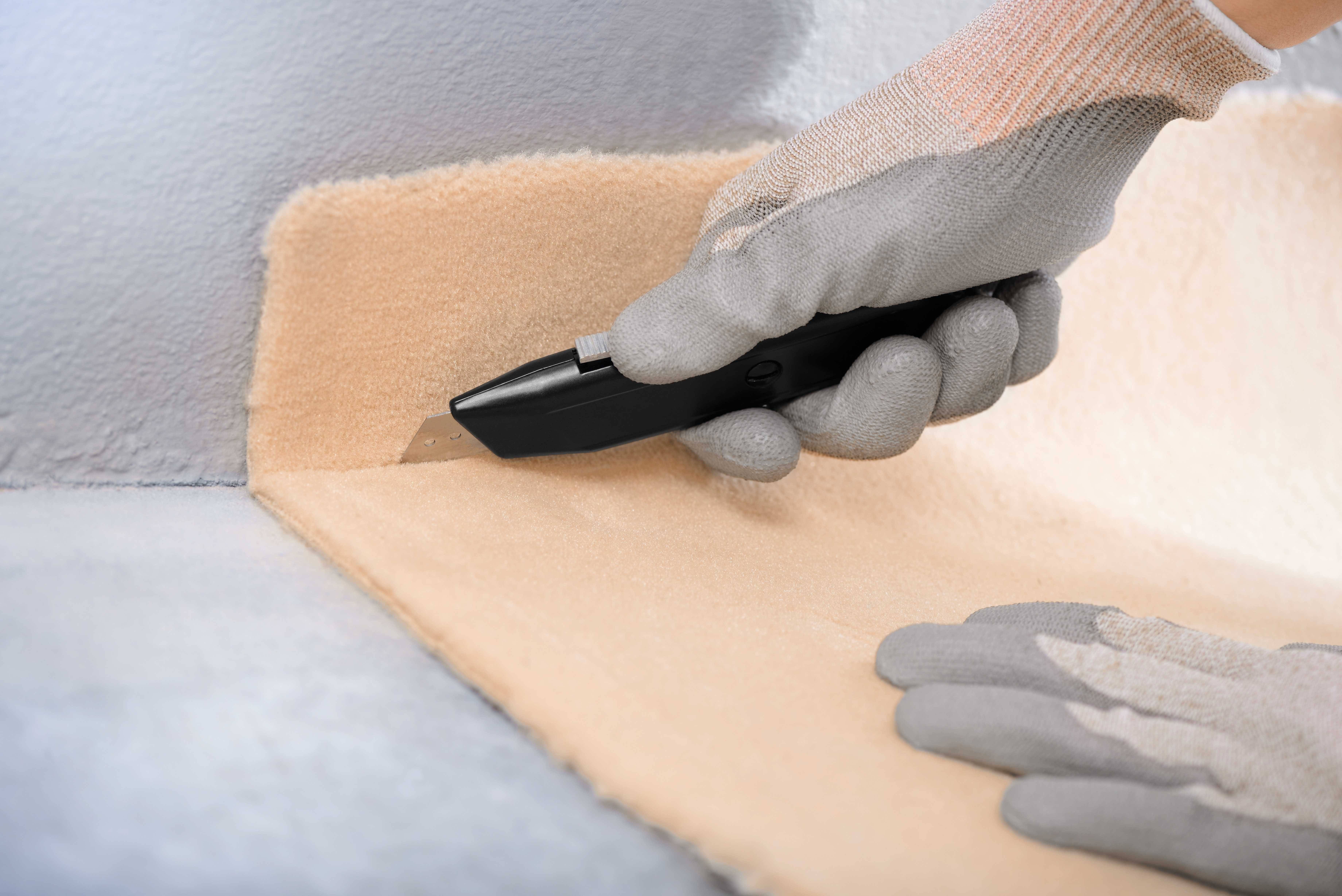 Eine behandschuhte Hand schneidet mit einem schwarzen Cutter-Messer beigefarbenen Teppich entlang einer Kante.