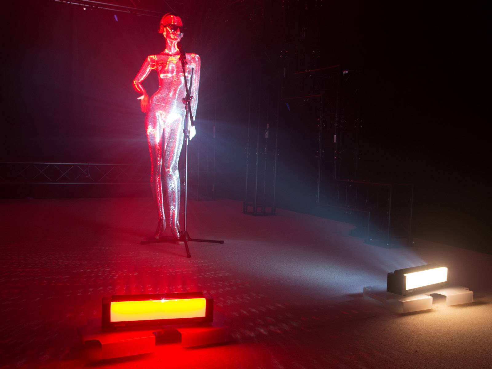 Eine glänzende, silberne Skulptur steht auf einer Bühne, beleuchtet von roten und weißen Scheinwerfern. Ein Mikrofon ist vor ihr platziert.
