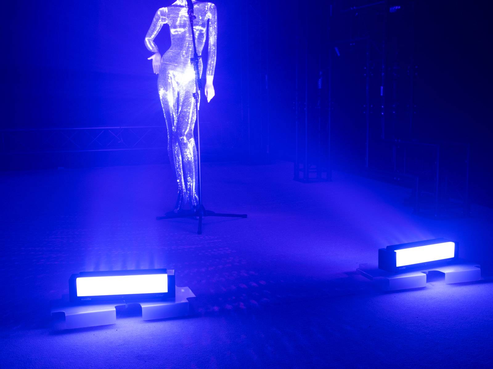 Eine glänzende Statue steht auf einer Bühne, umgeben von intensivem blauem Licht und scheinwerferähnlichen Lampen.