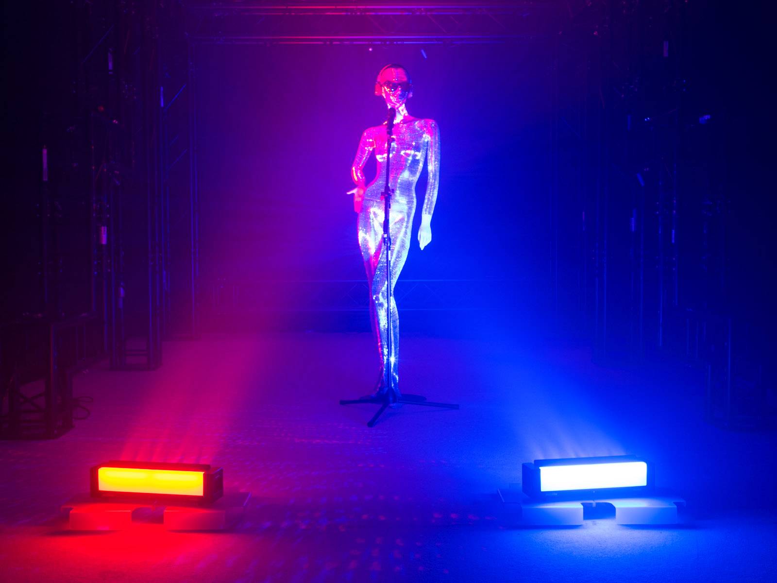 Eine glänzende, silberne Statue steht auf einer Bühne, beleuchtet von rotem und blauem Licht, mit einem Mikrofon vor sich.