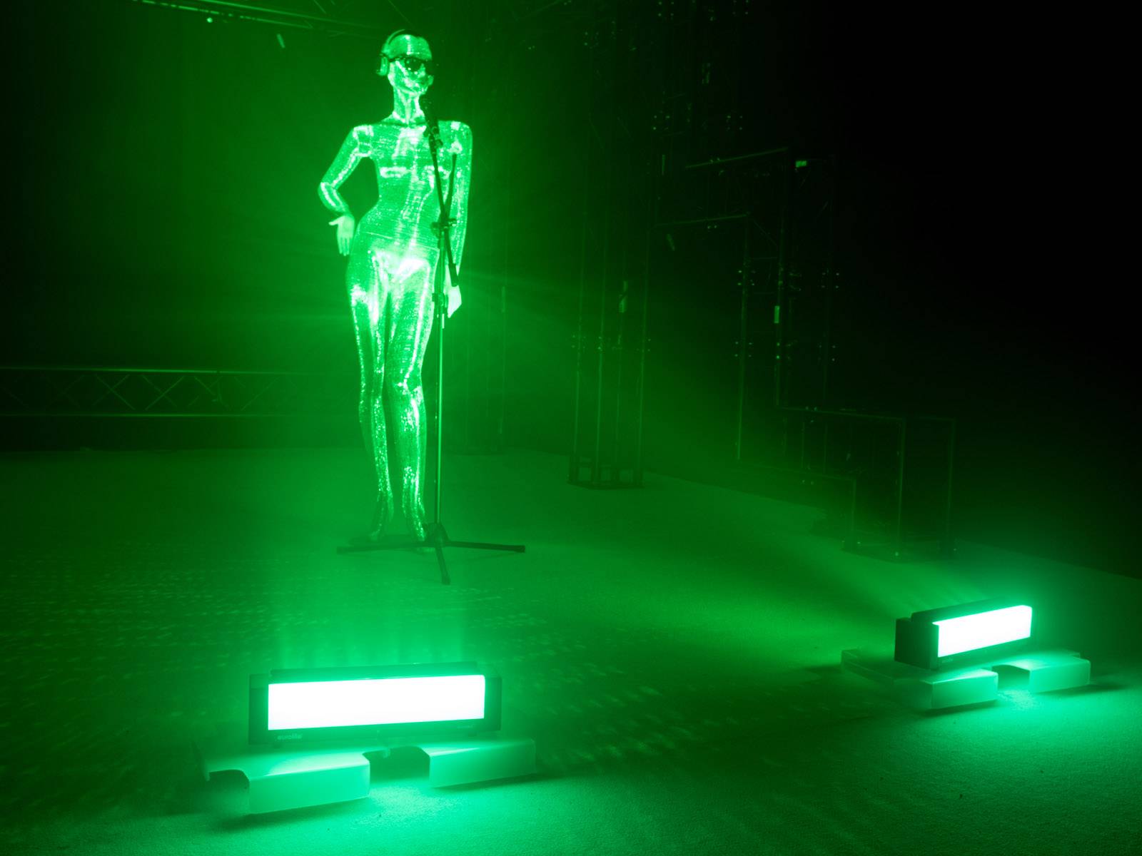Eine glänzende Figur steht in grünem Licht auf einer Bühne, möglicherweise während einer Performance oder Ausstellung.