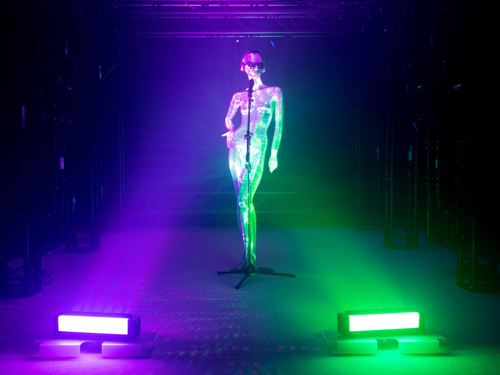 Eine glänzende, figurine Frau in der Mitte einer Bühne, beleuchtet von blau-violettem und grünem Licht, in einem dunklen Raum.