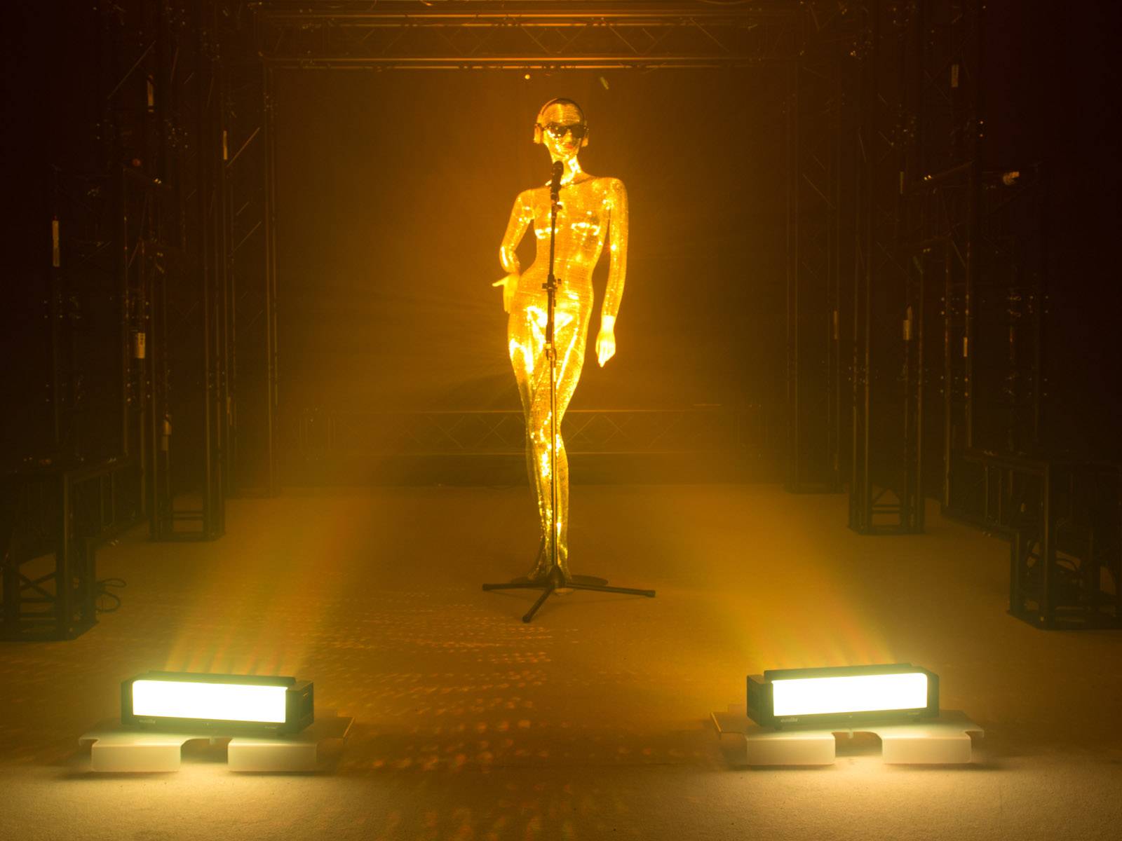 Eine goldene, menschliche Figur steht auf einer Bühne mit Mikrofon vor zwei leuchtenden Scheinwerfern, umgeben von dunklen Strukturen.