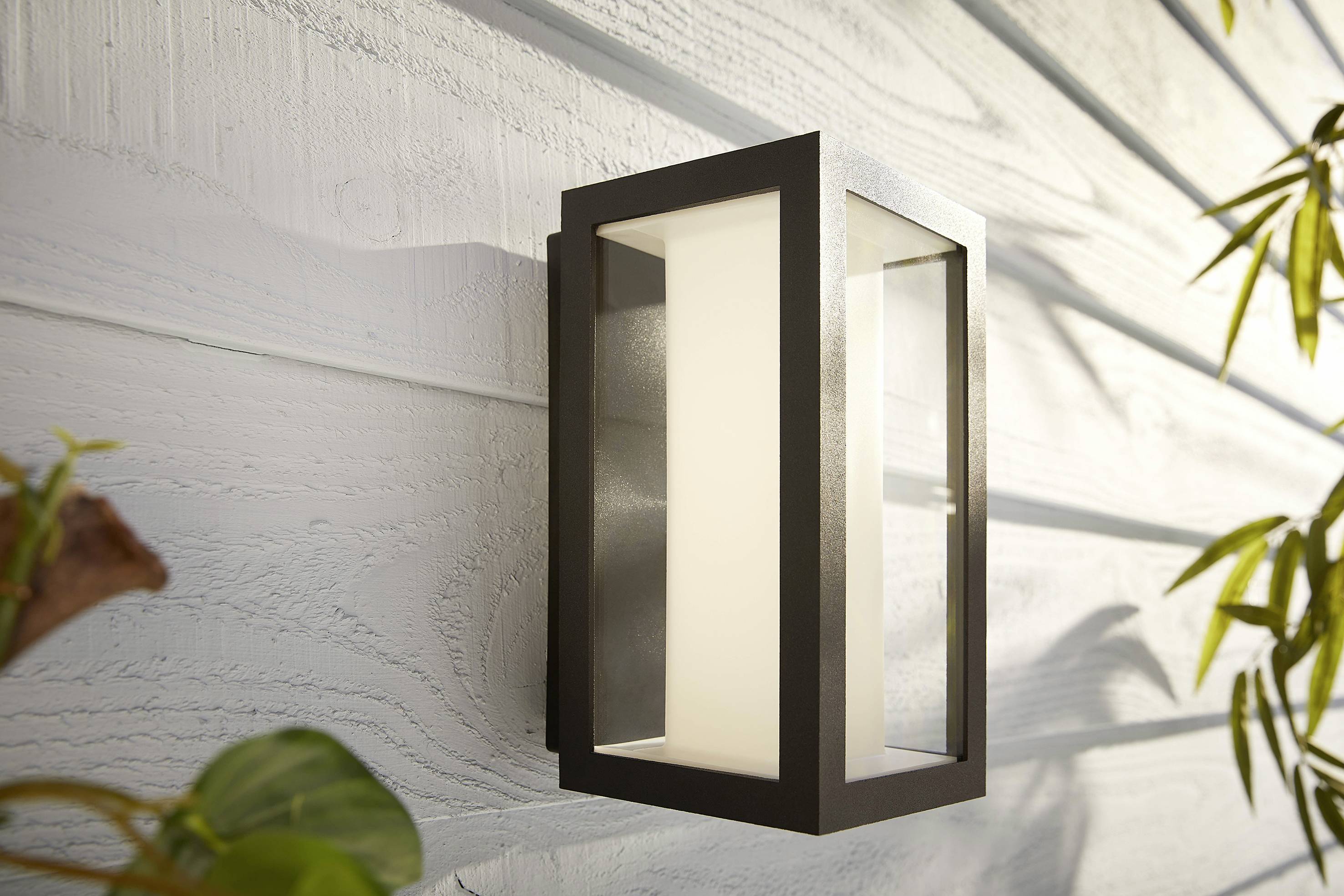 Wandleuchte in modernem, rechteckigem Design an einer hellen Außenwand, umgeben von Pflanzenblättern.