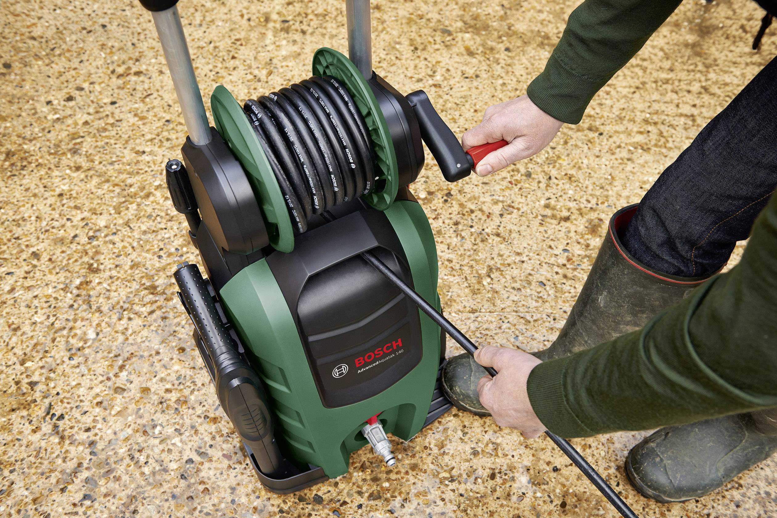 Ein Mann wickelt einen Gartenschlauch auf einem grünen Schlauchwagen von 'Bosch' auf. Er trägt Stiefel und steht auf einem Kiesboden.