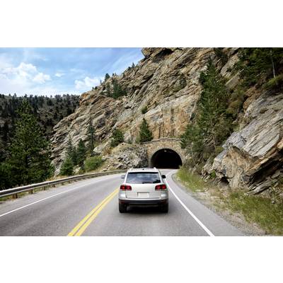Ein Auto fährt auf einer Straße durch eine felsige Landschaft, mit einem Tunnel im Hintergrund, umgeben von Bäumen und blauen Himmel.