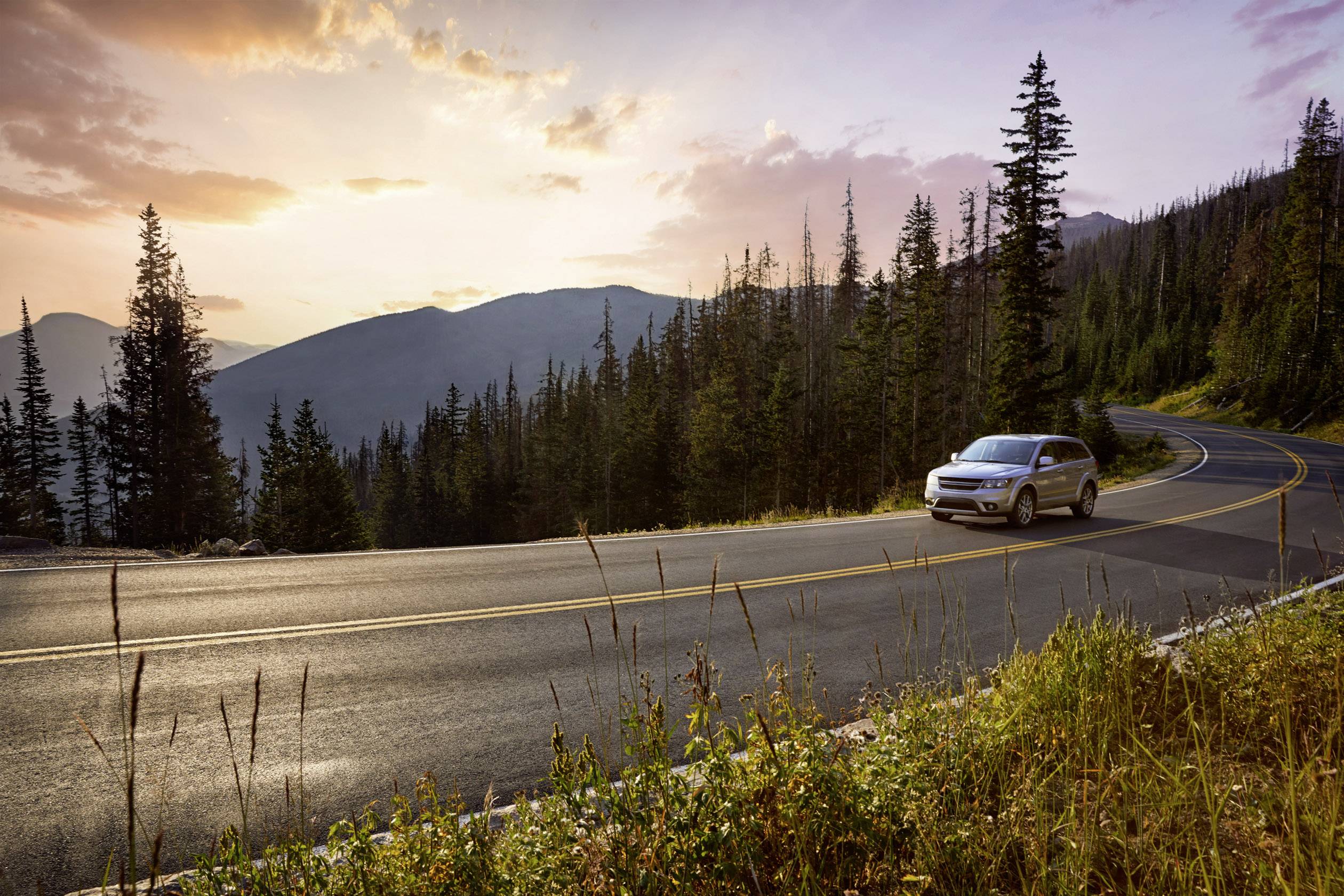 Auto fährt auf gewundener Straße durch bergige Waldlandschaft bei Sonnenuntergang, umgeben von hohen Bäumen und fernen Hügeln.