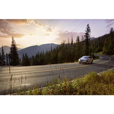 Auto fährt auf gewundener Straße durch bergige Waldlandschaft bei Sonnenuntergang, umgeben von hohen Bäumen und fernen Hügeln.