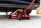 Ein roter Wagenheber hebt ein Auto in einer Garage an. Der Wagenheber befindet sich unter dem vorderen Teil des Fahrzeugs.