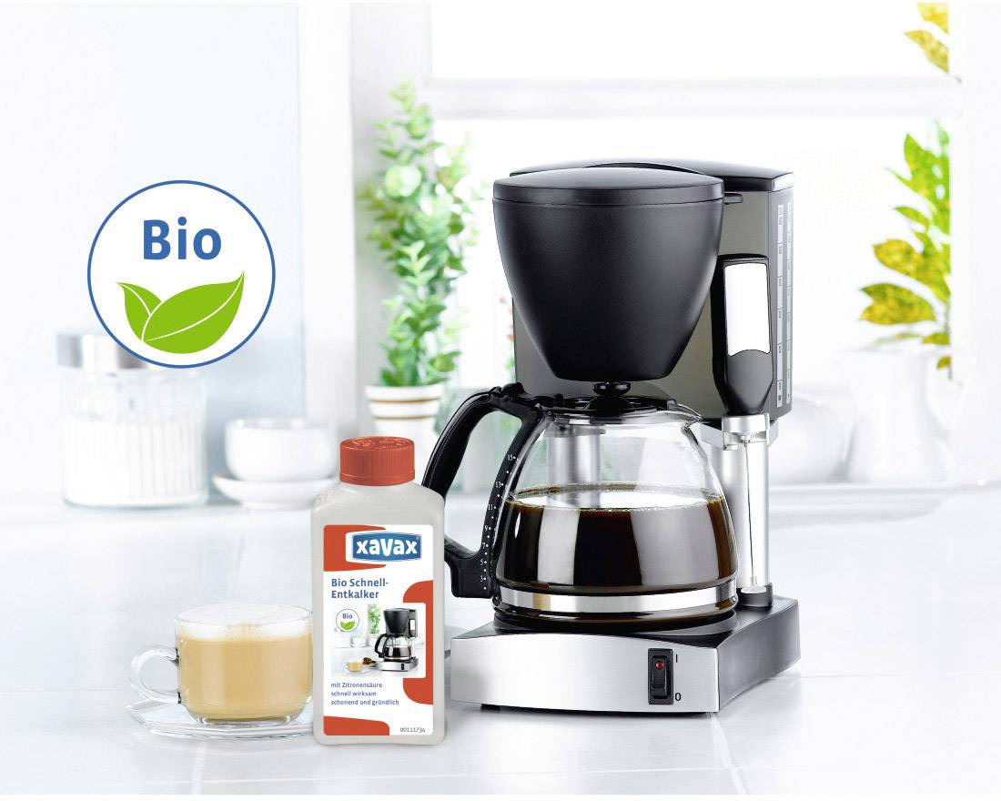 Eine Kaffeemaschine mit voller Glaskanne, daneben eine Packung Bio-Entkalker und eine Tasse Milchkaffee. Hintergrund: helle Küche.