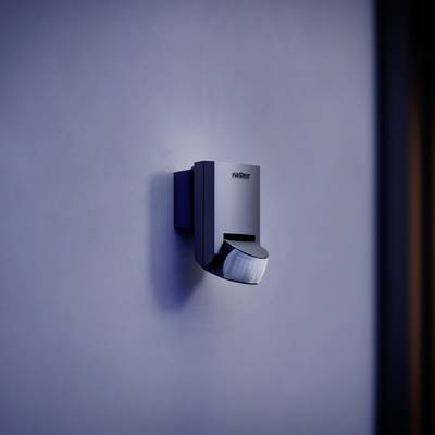 Ein Bewegungsmelder mit dunklem Gehäuse ist an einer blauen Wand montiert, beleuchtet von schwachem Licht.