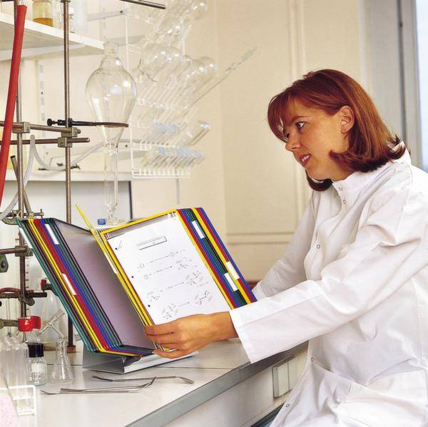 Eine Frau im Laborkittel überprüft ein Diagramm in einem Ordner an einem Chemielabor-Tisch mit verschiedenen Laborgeräten.