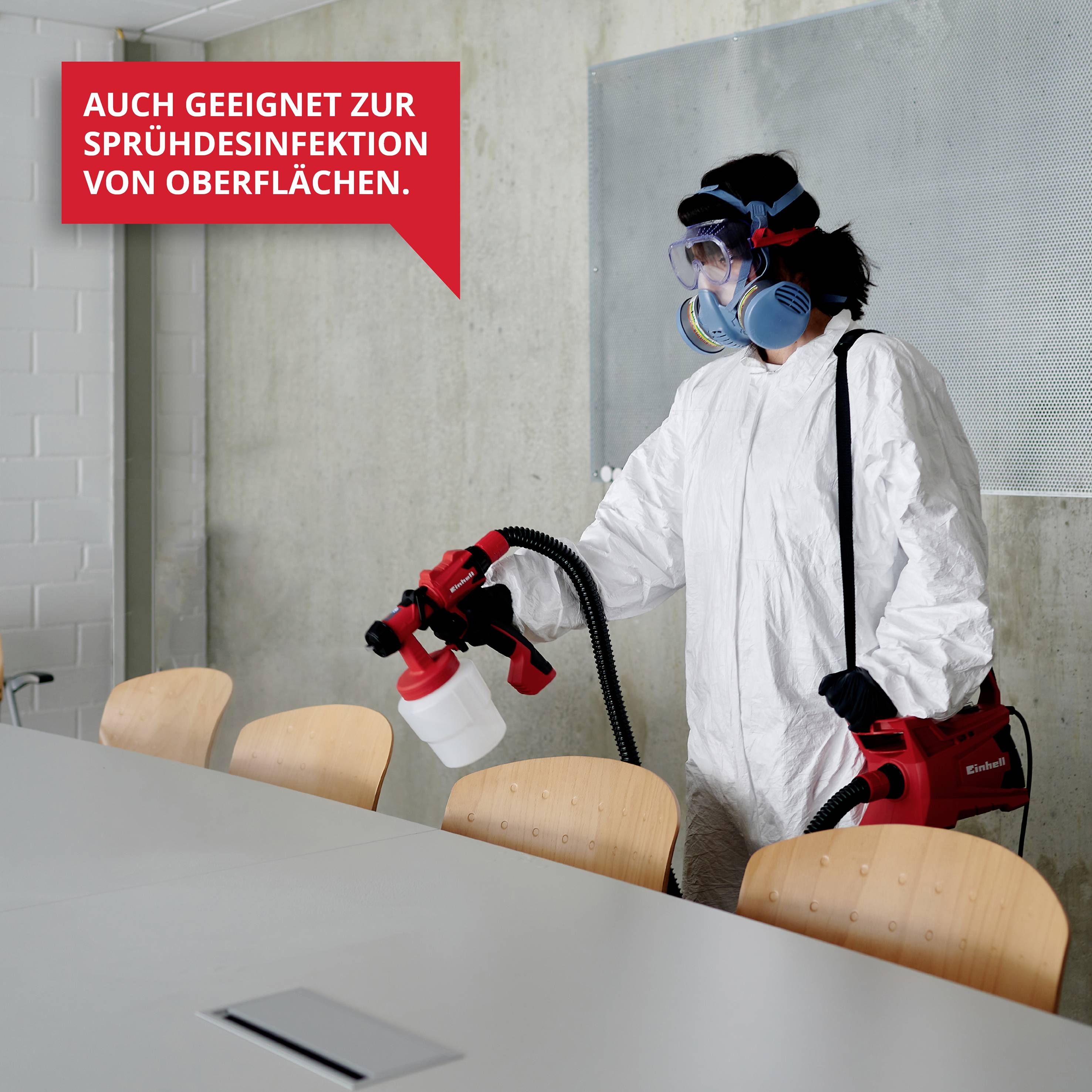 Person in Schutzanzug sprüht Desinfektionsmittel auf Oberflächen. Schild: 'Auch geeignet zur Sprühdesinfektion von Oberflächen'.