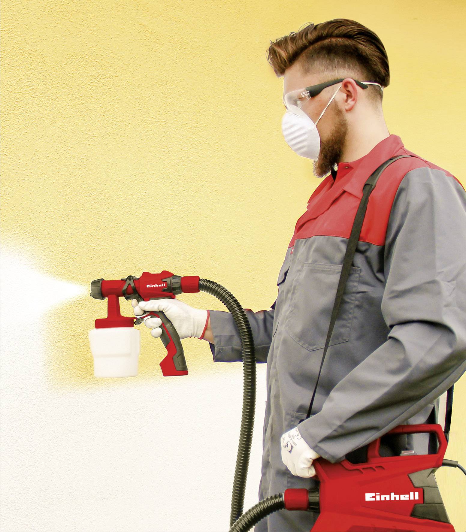 Ein Mann mit Schutzbrille und Maske sprüht gelbe Farbe mit einem Farbsprühsystem auf eine Wand. Er trägt graue Arbeitskleidung.