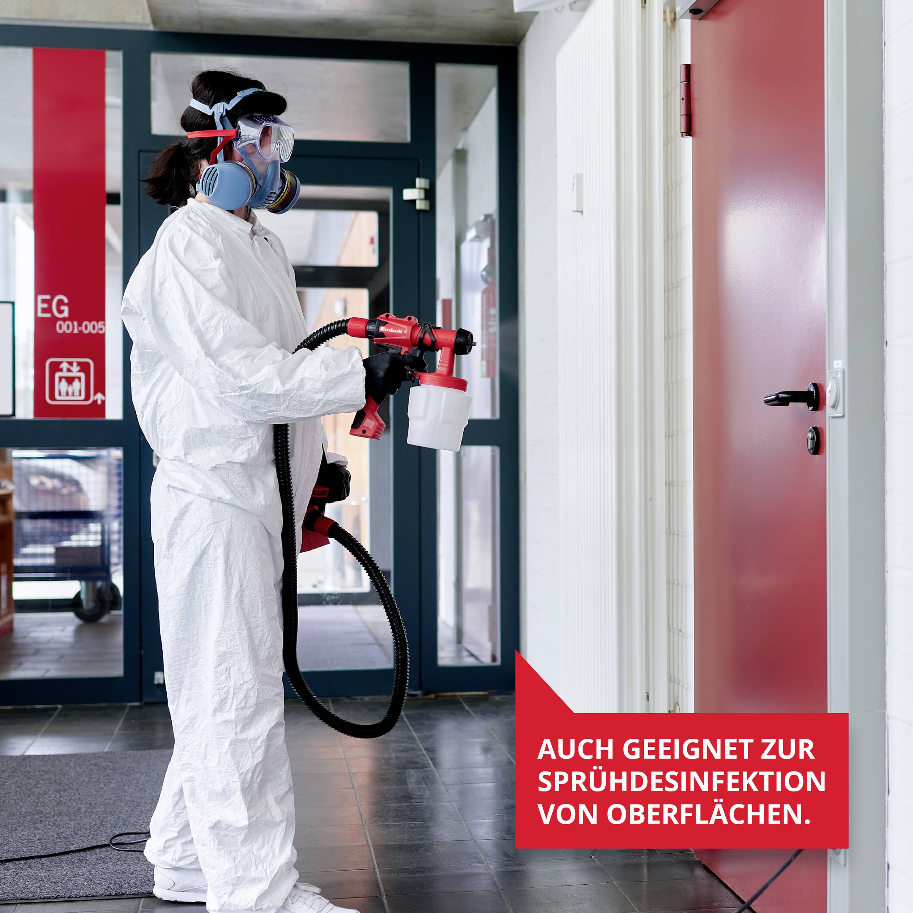 Person in Schutzanzug desinfiziert Tür in einem Gebäude mit einem Sprühgerät. Schild sagt: 'Auch geeignet zur Sprühdesinfektion von Oberflächen'.