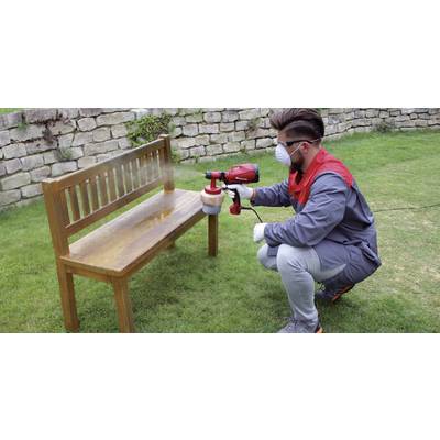 Eine Person lackiert eine Holzbank im Garten mit einer Spritzpistole. Sie trägt Schutzkleidung und eine Atemmaske.