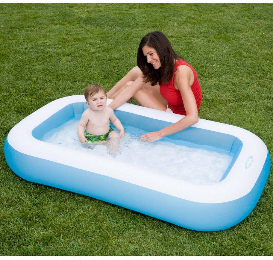 Eine Frau in einem roten Oberteil spielt in einem blauen aufblasbaren Kinderpool im Garten mit einem Kleinkind, das im Wasser sitzt.