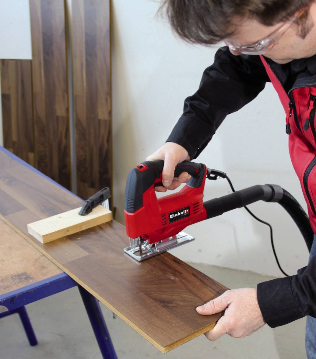 Eine Person schneidet mit einer Stichsäge ein Holzbrett auf einem Tisch. Späne werden aufgesaugt. Im Hintergrund stehen weitere Bretter.