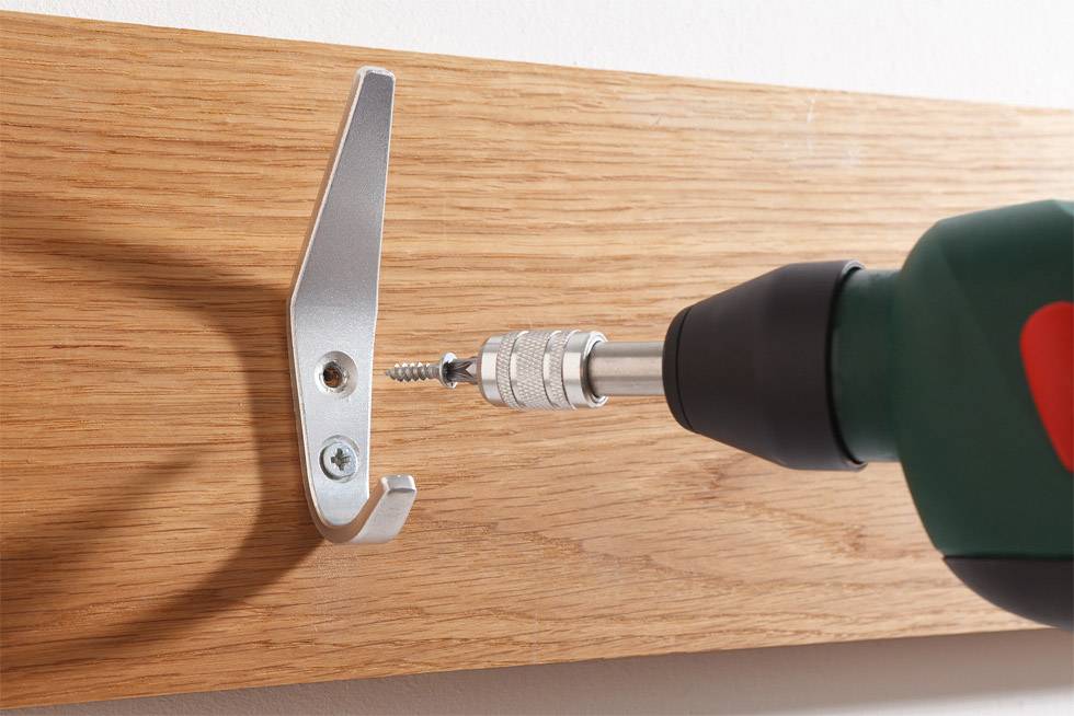 Eine grüne Bohrmaschine befestigt einen silbernen Haken mit einer Schraube auf einem Holzbrett an der Wand.