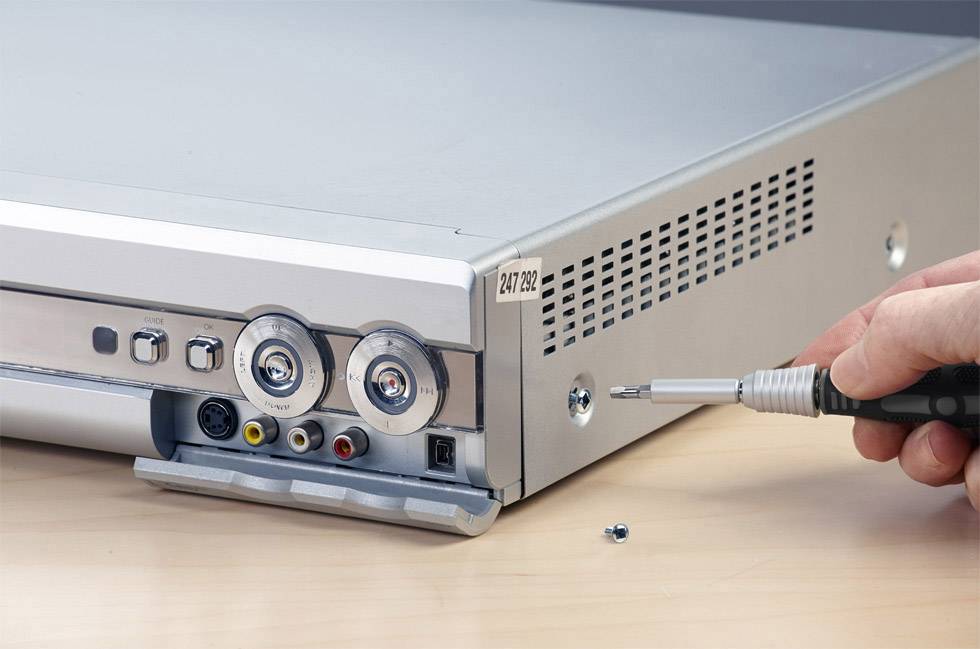 Eine Person öffnet das Gehäuse eines Videorecorders, indem sie mit einem Schraubendreher eine Schraube auf der rechten Seite löst.