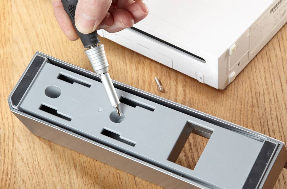 Eine Hand hält einen Schraubendreher, der einen grauen Kunststoff-Gehäusedeckel neben einem weißen elektronischen Gerät entfernt.