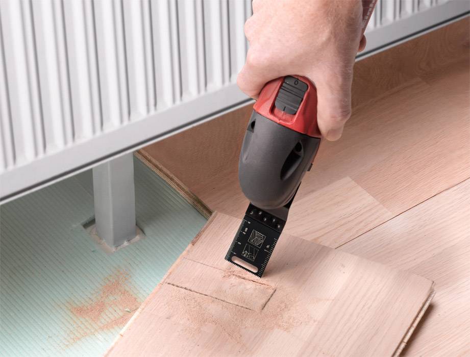 Eine Hand hält ein elektrisches Multifunktionswerkzeug, das ein rechteckiges Stück Holz aus einem Bodenbelag schneidet.