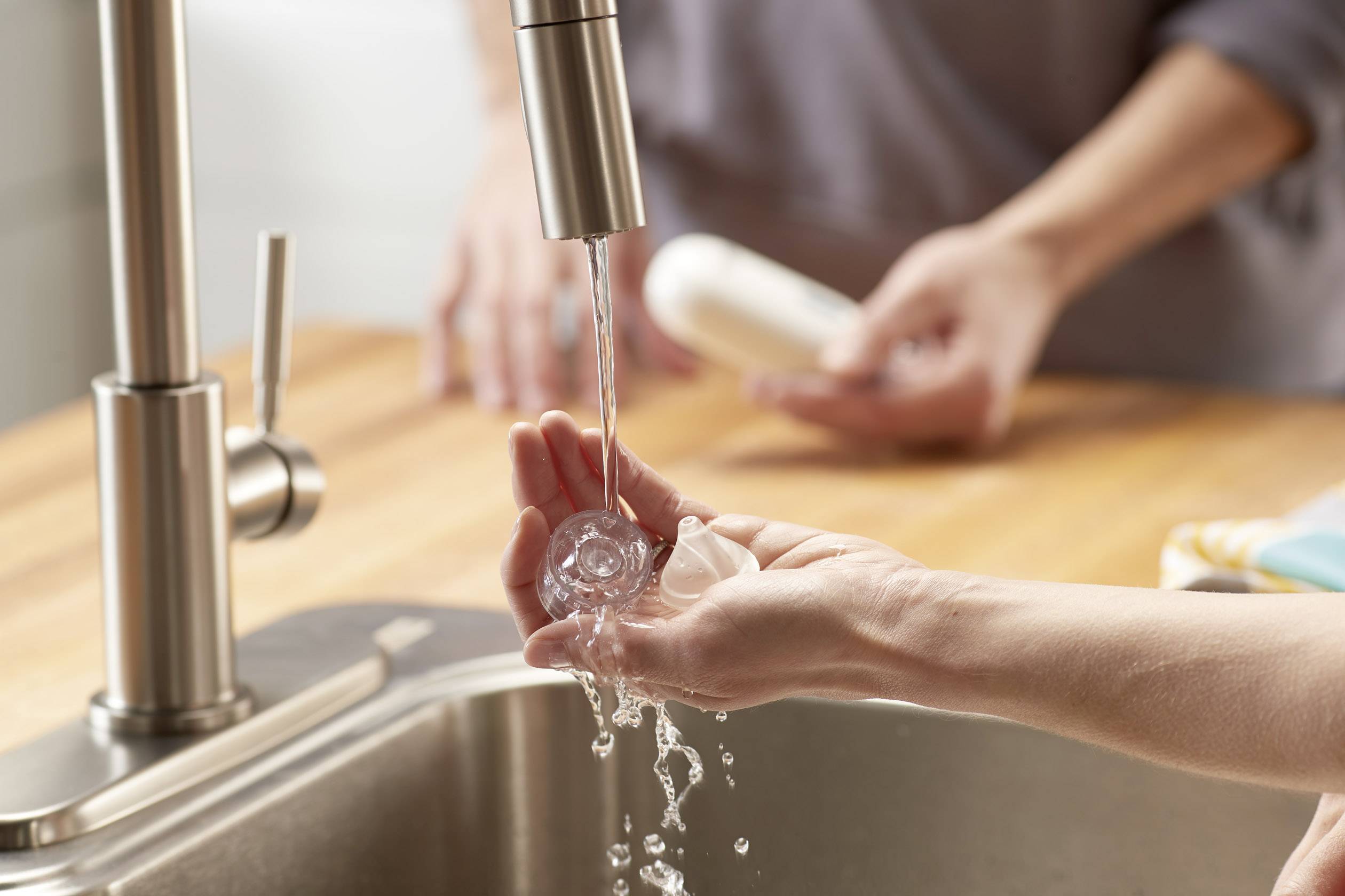 Nahaufnahme von Händen, die unter einem Wasserhahn in einer Küche Gemüse waschen, mit einer zweiten Person im Hintergrund.