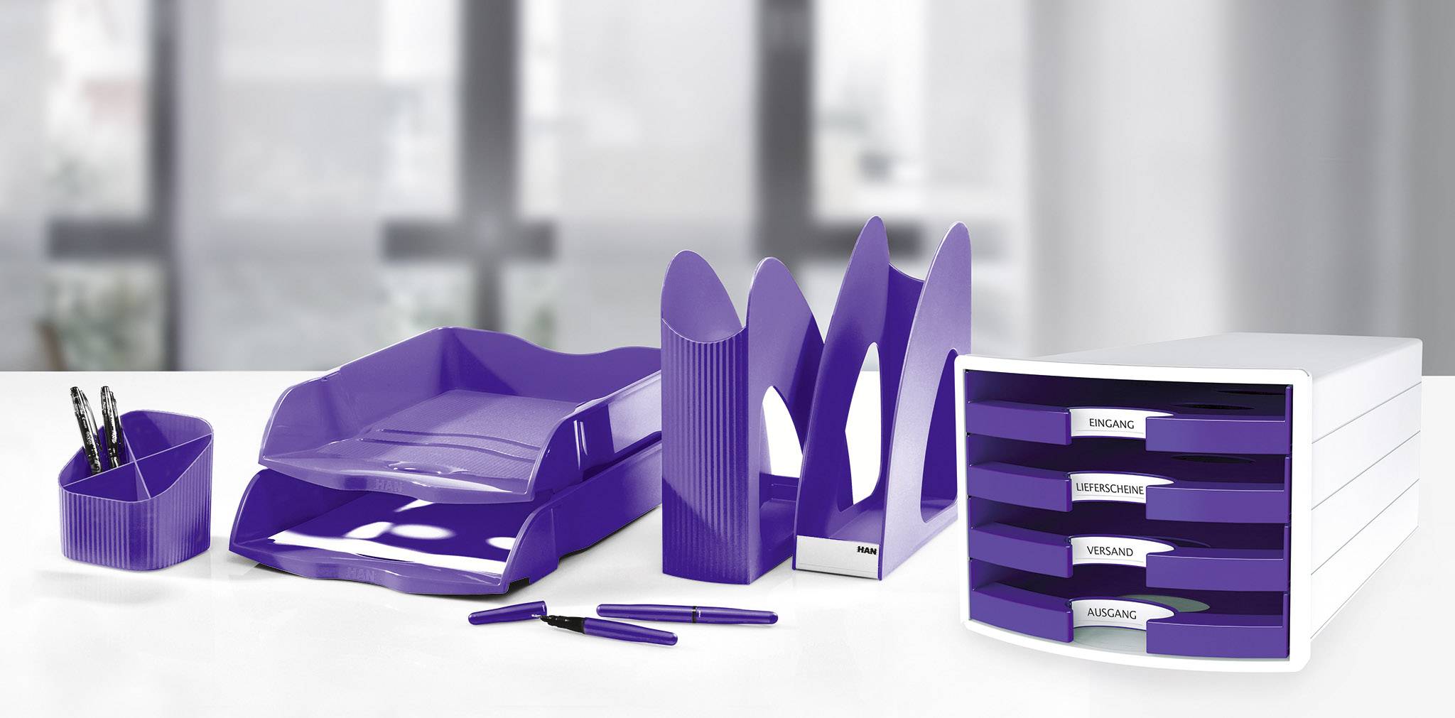 Lila Büroorganisation: Briefablagen, Stifthalter, Stehsammler und Schubladenbox auf einem weißen Tisch. Büroartikel in modernem Design.