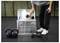 Eine Person legt eine Hantel in eine Metalllagerkiste in einer Sporthallumgebung, während Kettlebells und eine weitere Hantel in der Nähe auf dem Boden liegen.