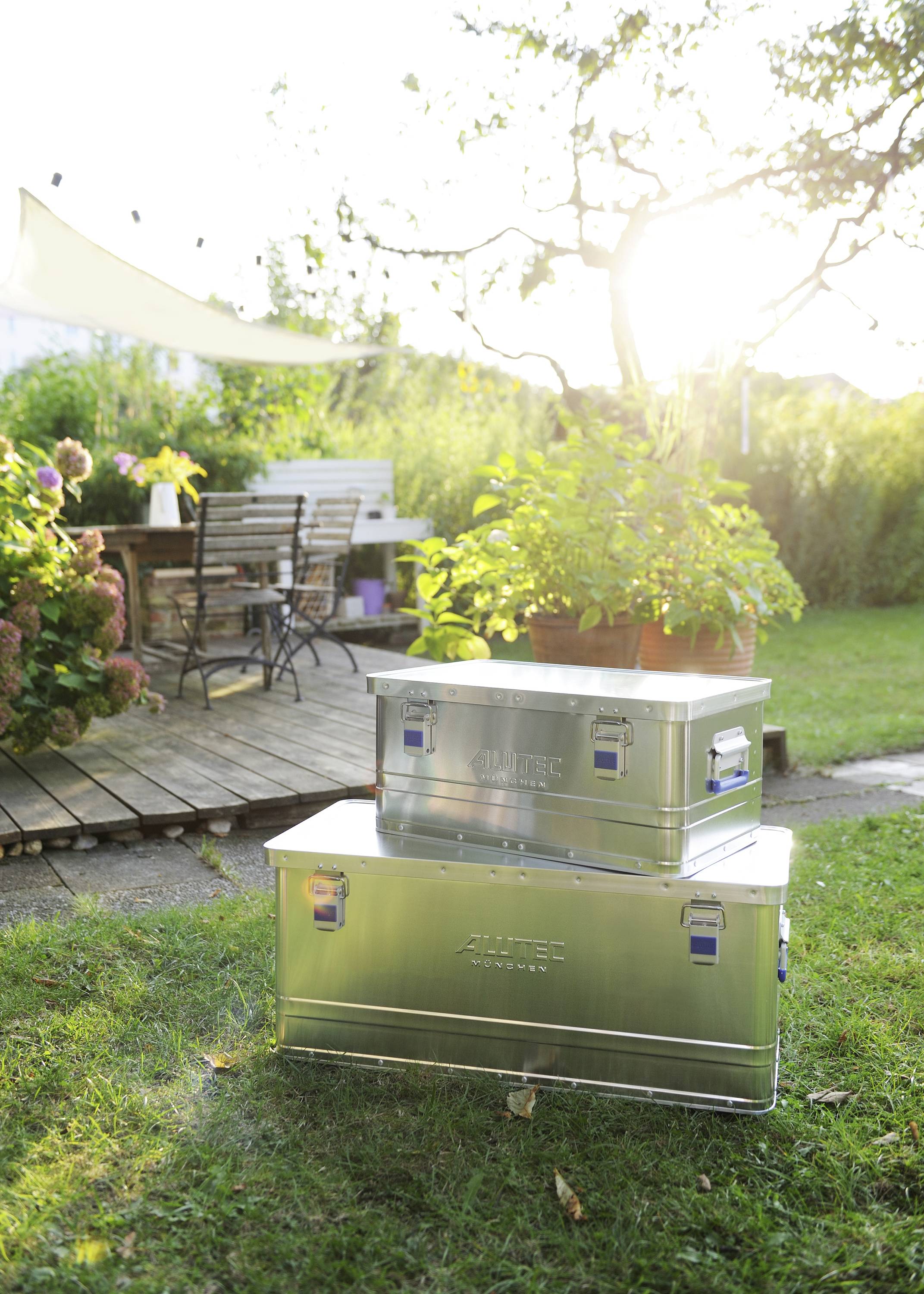 Zwei Metallkisten im Garten, eine mit Topfpflanze darauf. Im Hintergrund ein Holztisch mit Stühlen auf einem Holzdeck unter Bäumen.